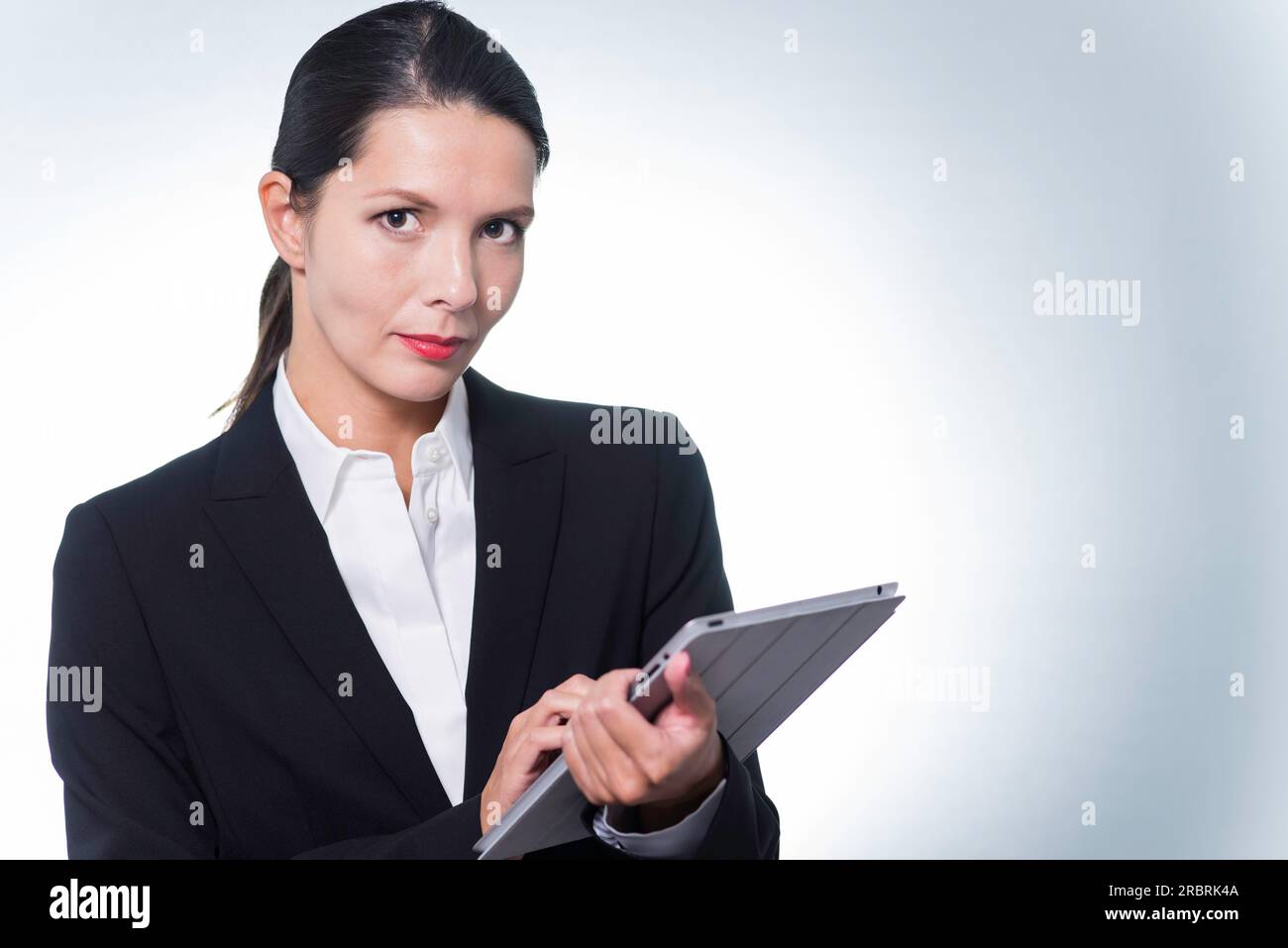 Confident business manageress working on a tablet computer as she stands looking at the camera with a serious thoughtful expression, studio portrait Stock Photo