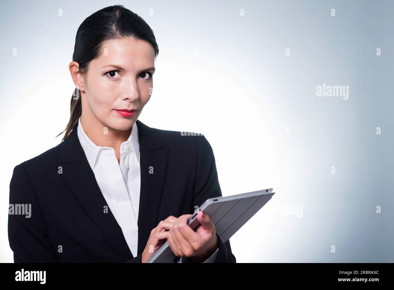 Confident business manageress working on a tablet computer as she stands looking at the camera with a serious thoughtful expression, studio portrait Stock Photo