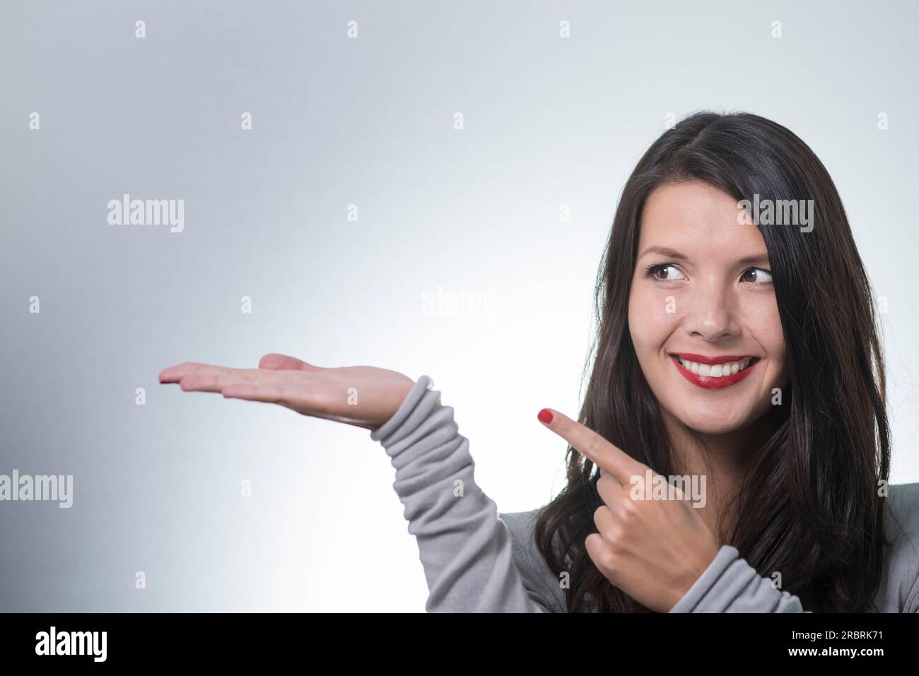 Beautiful young woman with a lovely friendly smile standing with an outstretched empty palm for your product placement pointing to it with her other Stock Photo