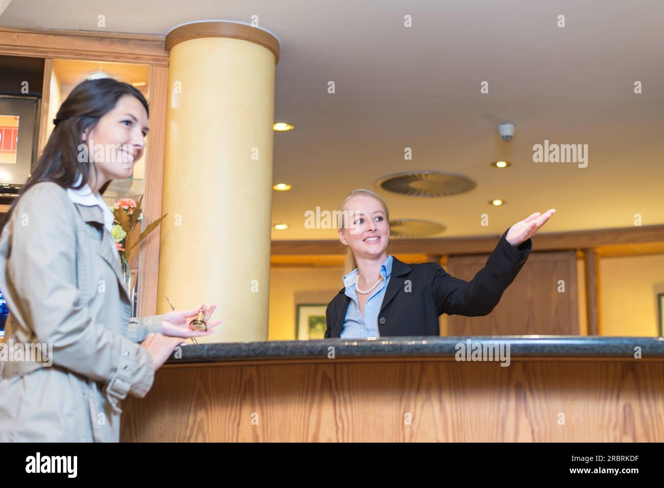 Low angle view of a beautiful friendly smiling receptionist behind the service desk in a hotel lobby helping an attractive female guest indicating Stock Photo