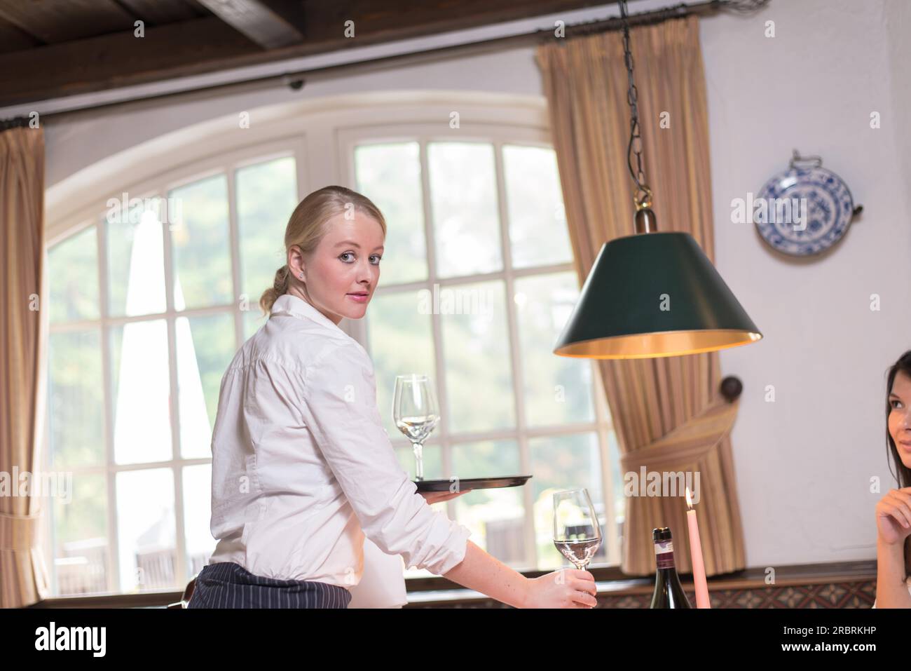Low angle view of a beautiful young waitress serving drinks in a restaurant pausing to turn round and look at the camera as she stretches out her Stock Photo