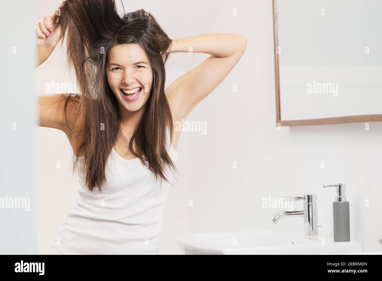 Woman yelling in frustration as she brushes her hair standing in the bathroom tugging at her unruly locks and tangles Stock Photo