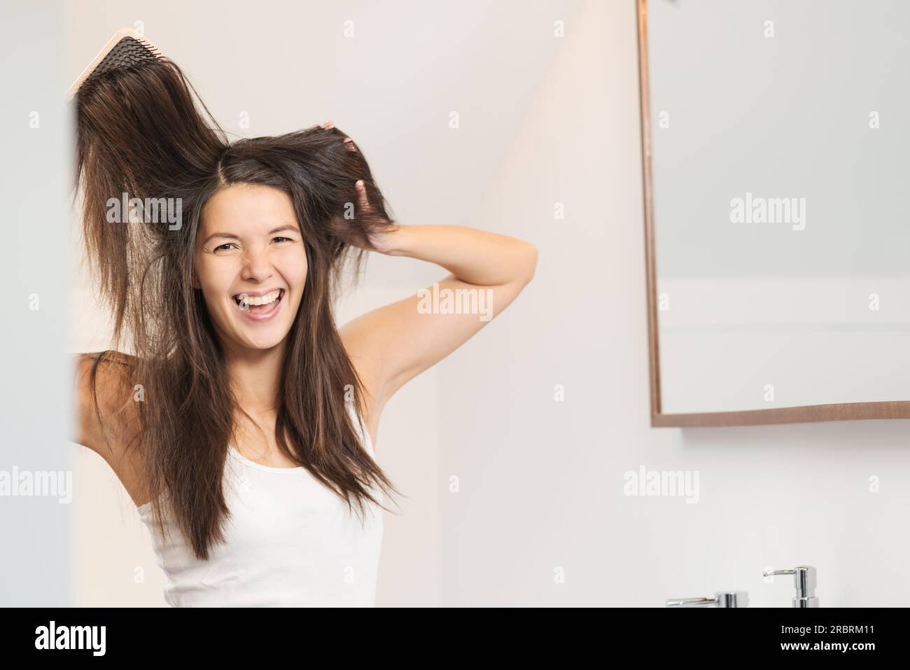 Woman yelling in frustration as she brushes her hair standing in the bathroom tugging at her unruly locks and tangles Stock Photo