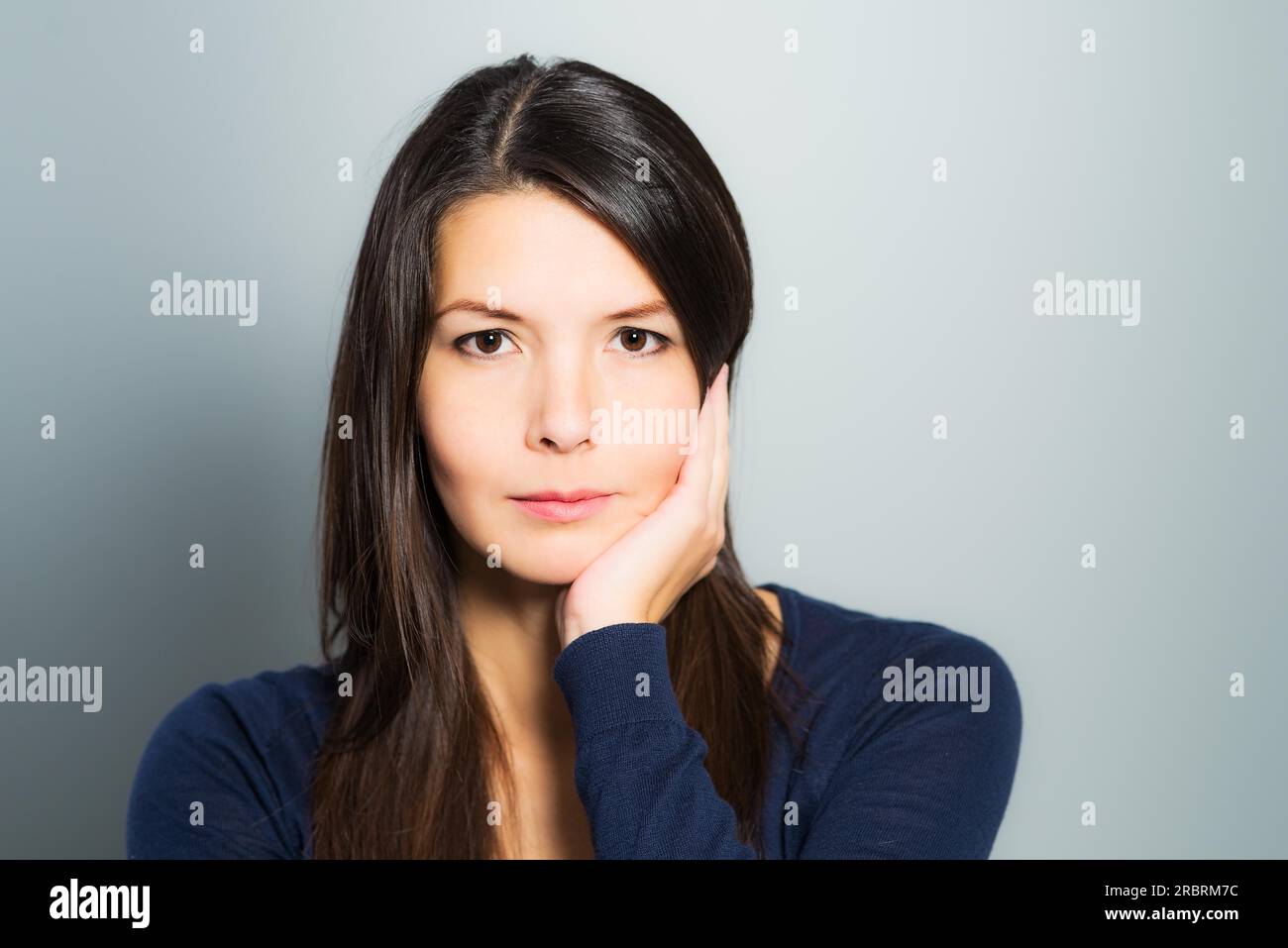 Thoughtful attractive woman with a serene face and enigmatic smile standing with her hand to her chin looking pensively at the camera Stock Photo