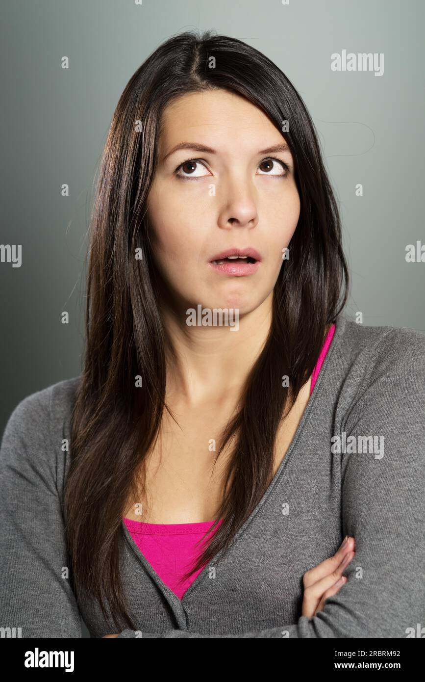Attractive woman with a sceptical bemused expression standing with folded arms pulling a wry face as she looks upwards into the air Stock Photo