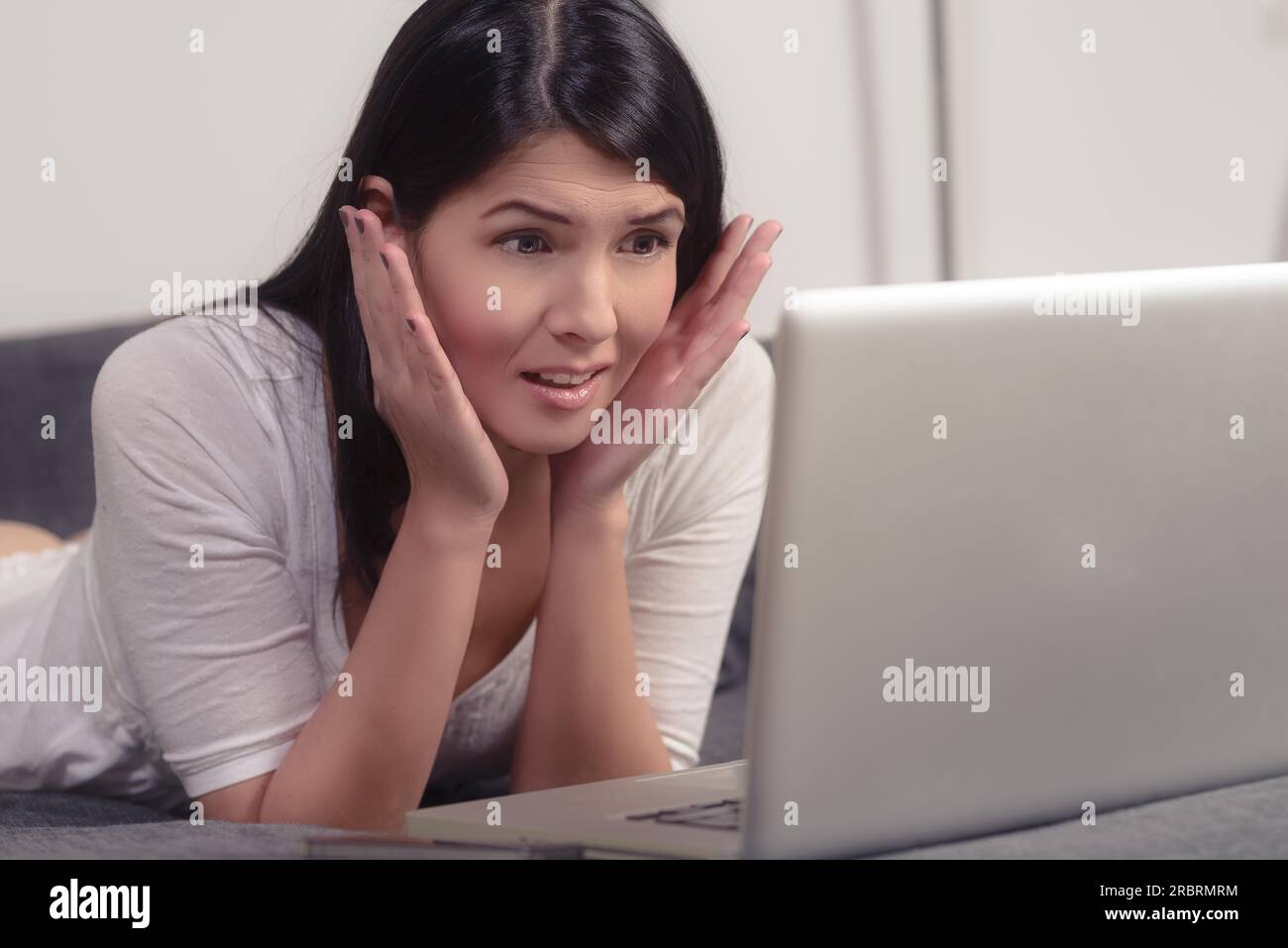 Beautiful young woman looking at her laptop computer with a horrified expression and her hands cupping her face as she reads something upsetting on Stock Photo