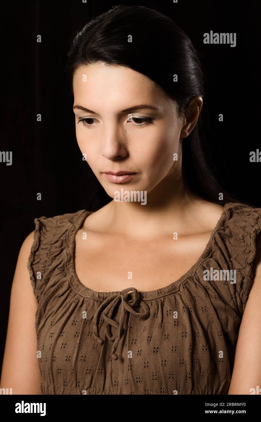 Melancholy depressed young woman with a serious expression and downcast eyes standing thinking in a deep contemplation with her hand to her chin Stock Photo