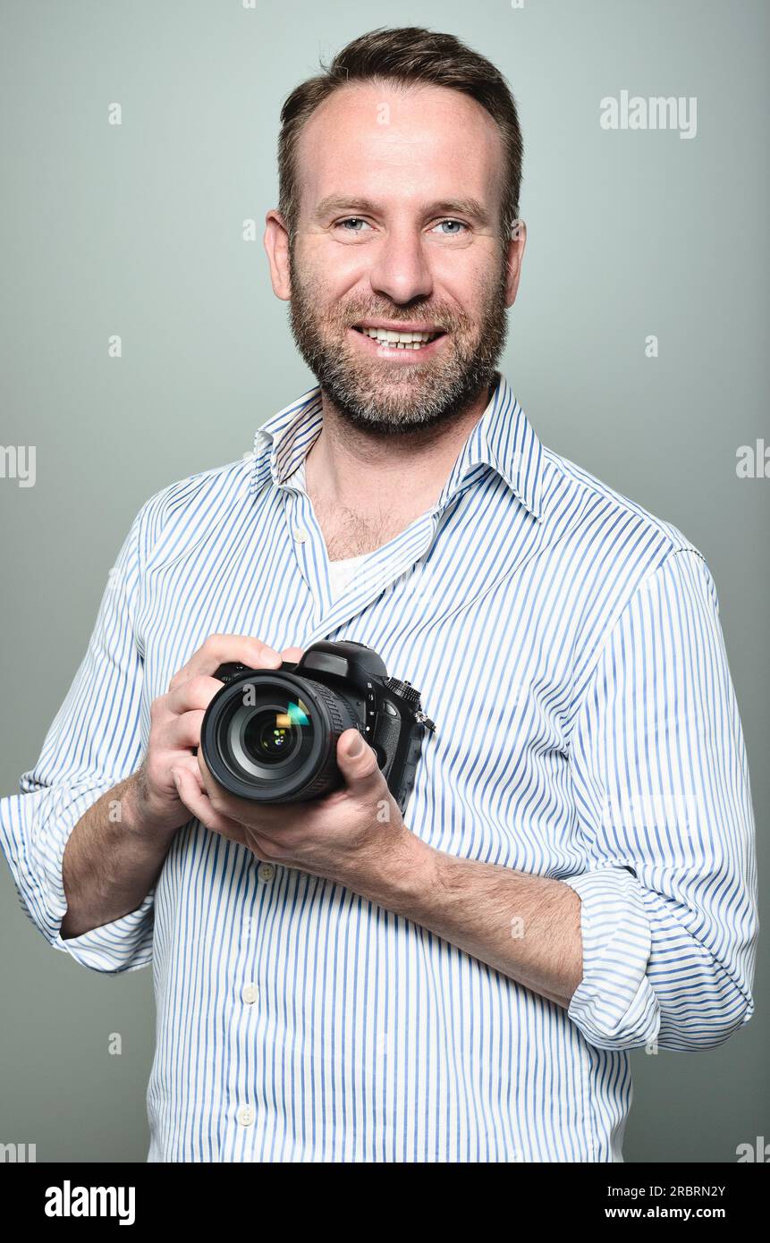 Handsome photographer with a friendly smile standing holding his camera  laughing at the camera, upper body portrait in relaxed pose on grey Stock  Photo - Alamy