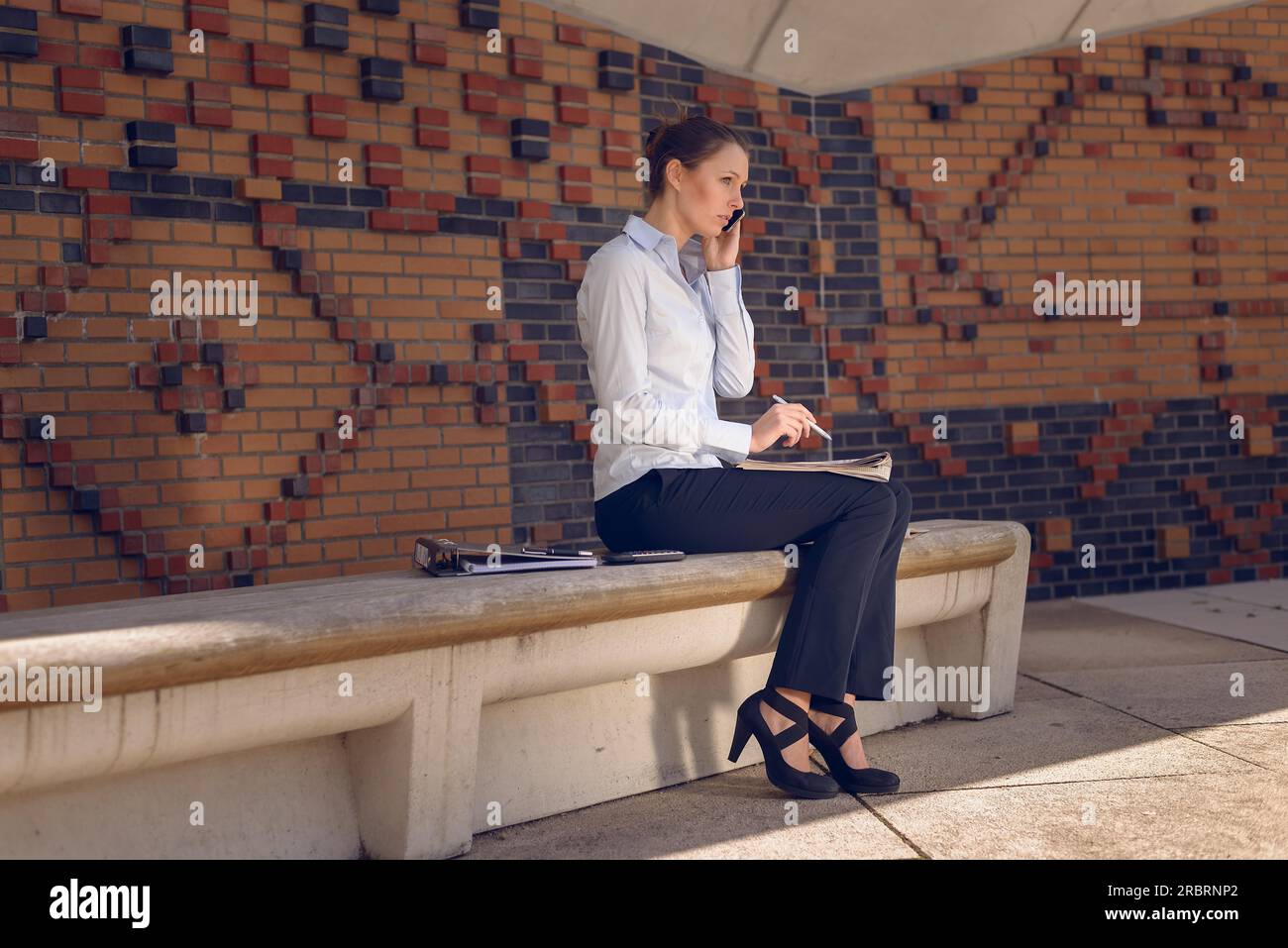 Woman doing business in a commercial foyer sitting on a wall mounted bench against patterned brickwork talking on her mobile phone discussing her Stock Photo