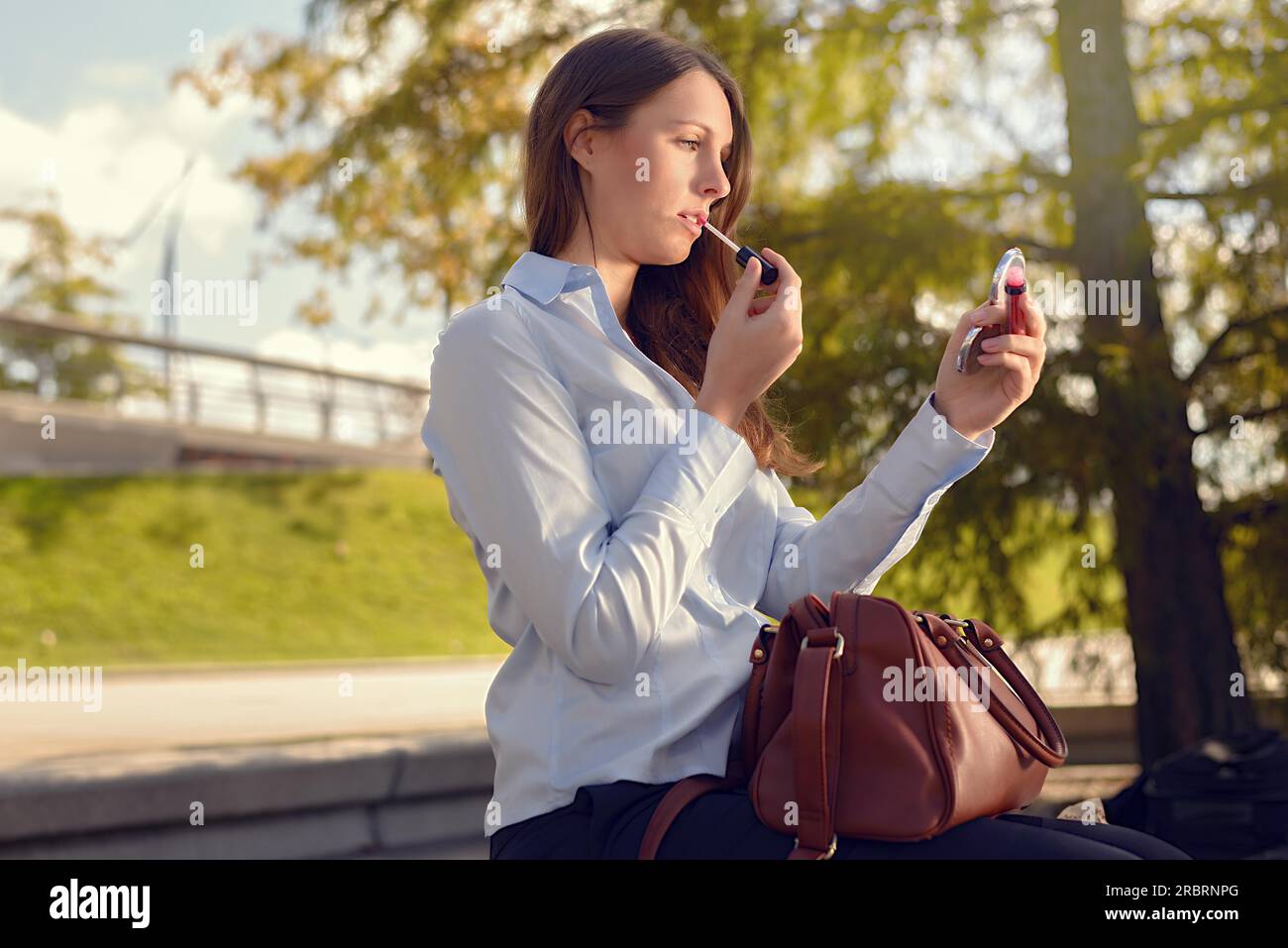 Attractive young woman refreshing her makeup in the street applying lipstick with the use of a small handheld mirror from her large handbag Stock Photo