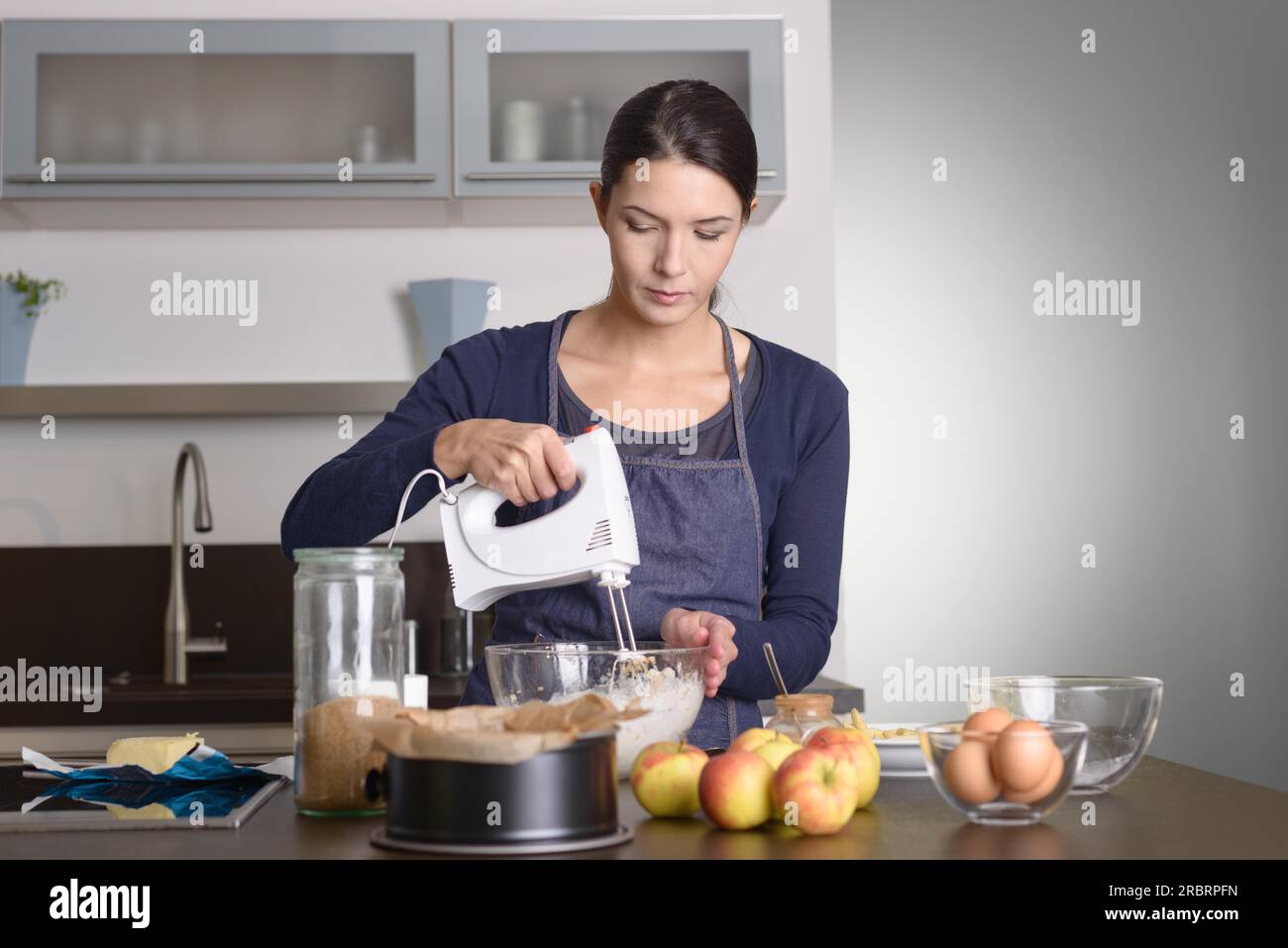 Young woman baking an apple pie in the kitchen standing at the counter in her apron using a handheld mixer to whisk the fresh ingredients in a glass Stock Photo