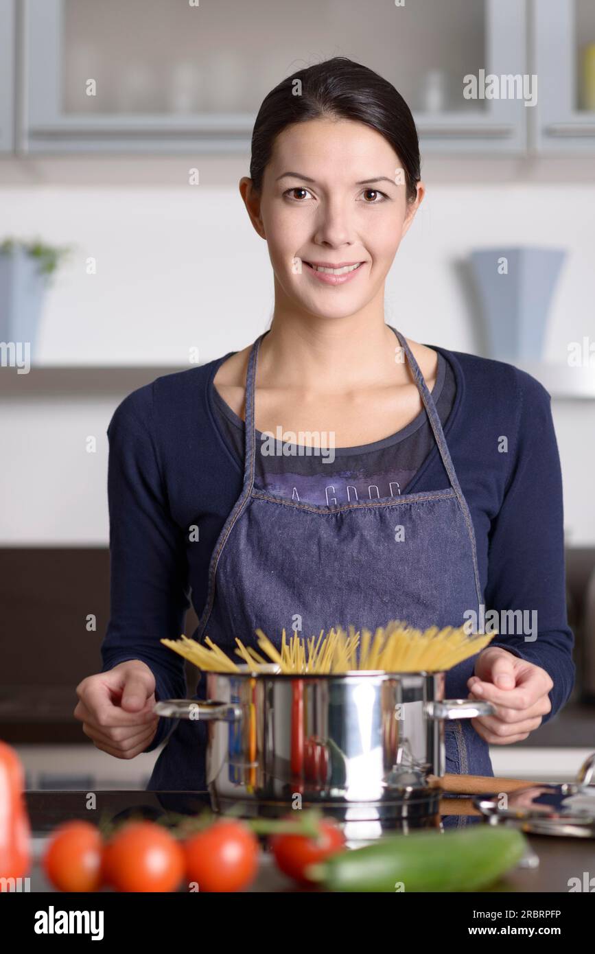 Young housewife preparing a healthy Italian pasta standing at the stove with a pot full of spaghetti with fresh vegetables in the foreground smiling Stock Photo