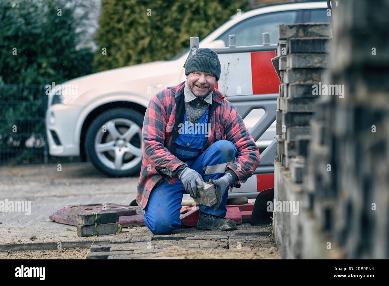 Cheerful workman dressed in warm winter clothing kneeling at the roadside laying paving bricks in his overalls looking at te camera with a beaming Stock Photo