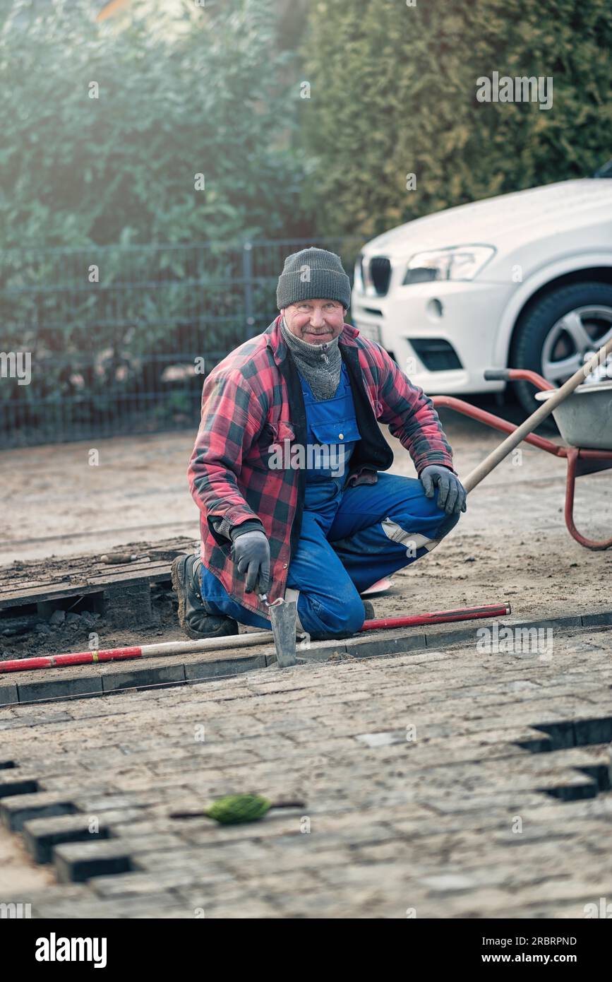 Cheerful workman dressed in warm winter clothing kneeling at the roadside laying paving bricks in his overalls looking at te camera with a beaming Stock Photo