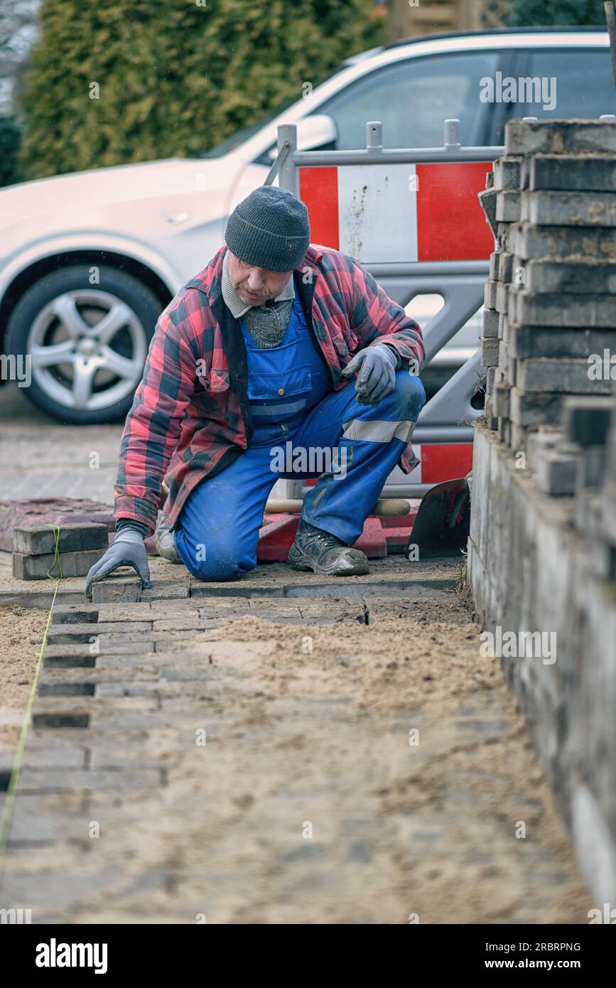 Concentrated workman dressed in warm winter clothing kneeling at the roadside laying paving bricks in his overalls looking at te camera with a Stock Photo