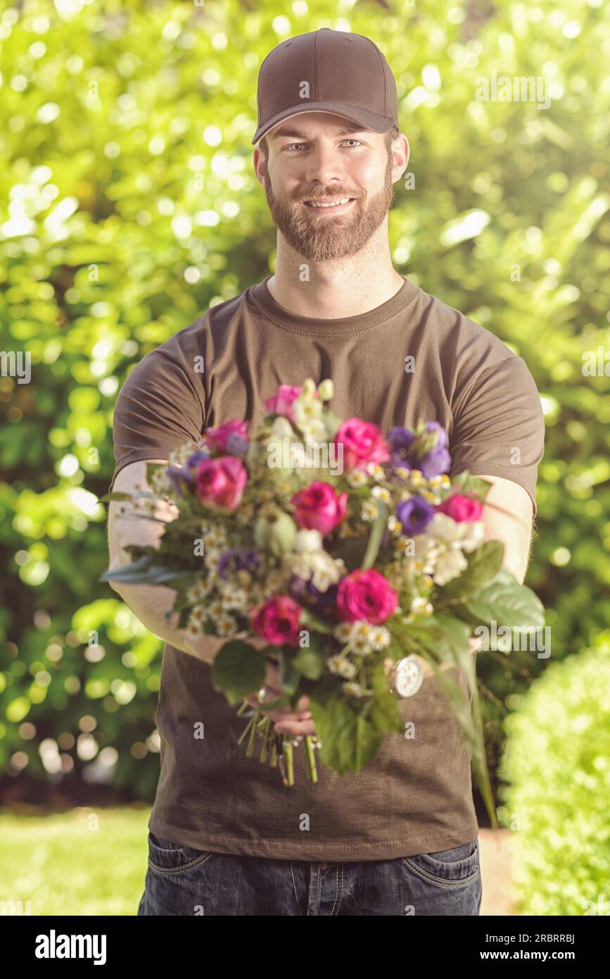 Three quarter length front view of smiling bearded 20s man wearing brown baseball cap, brown t-shirt and jeans holding bunch of flowers Stock Photo