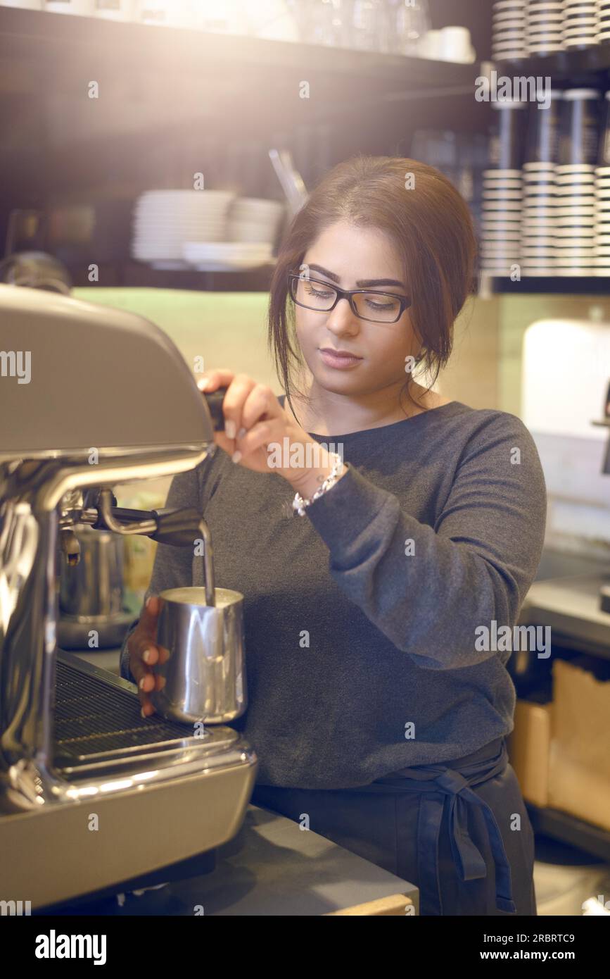 Female barista or waitress making a cup of cappuccino in a coffee house or cafeteria pouring the hot frothy milk into the coffee cup Stock Photo