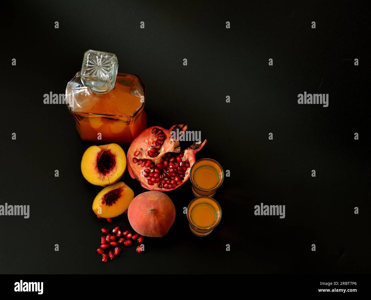 Two glasses and a glass decanter with homemade liqueur on a black background, next to pieces of a ripe peach and a broken pomegranate. Top view, flat Stock Photo
