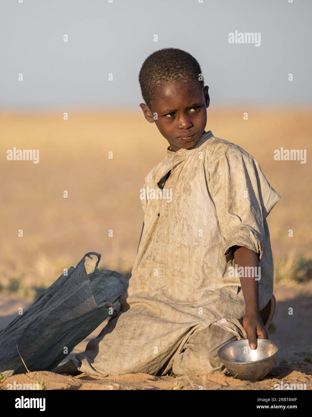 Young boy, clad in a weathered robe, humbly kneels on the steppe ground ...