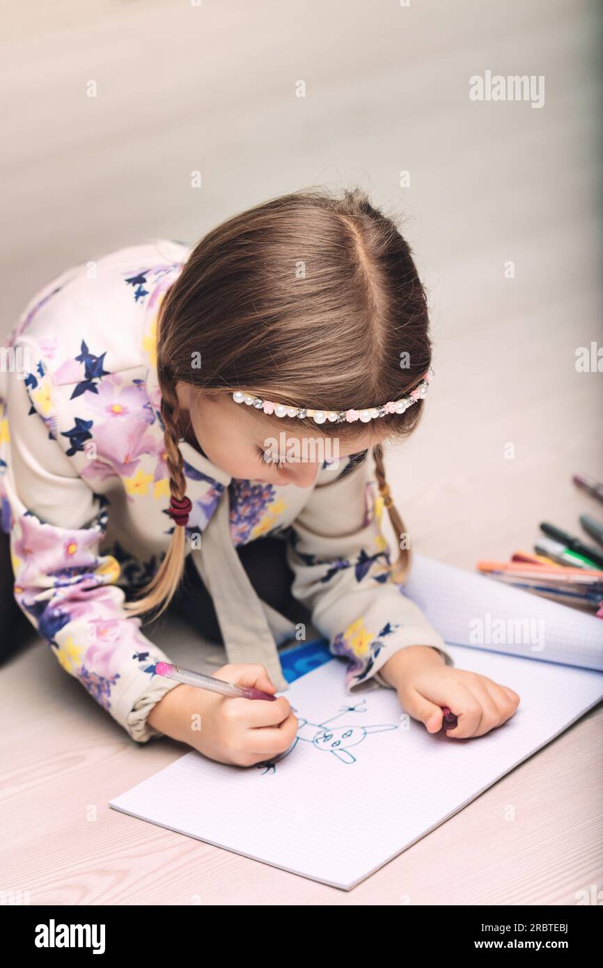 A small girl, focused on drawing and coloring, creates imaginary creatures and stories. Lying belly-down on the floor, she's absorbed in her play, a k Stock Photo