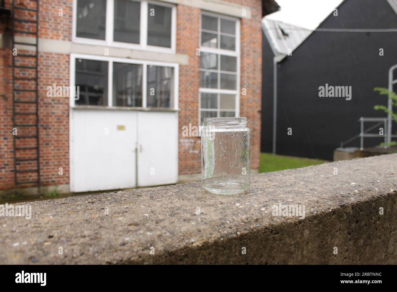 Old jar used as ash tray covered in rain drops while standing on a concrete wall. An abandoned industrial brick building in the background. Stock Photo
