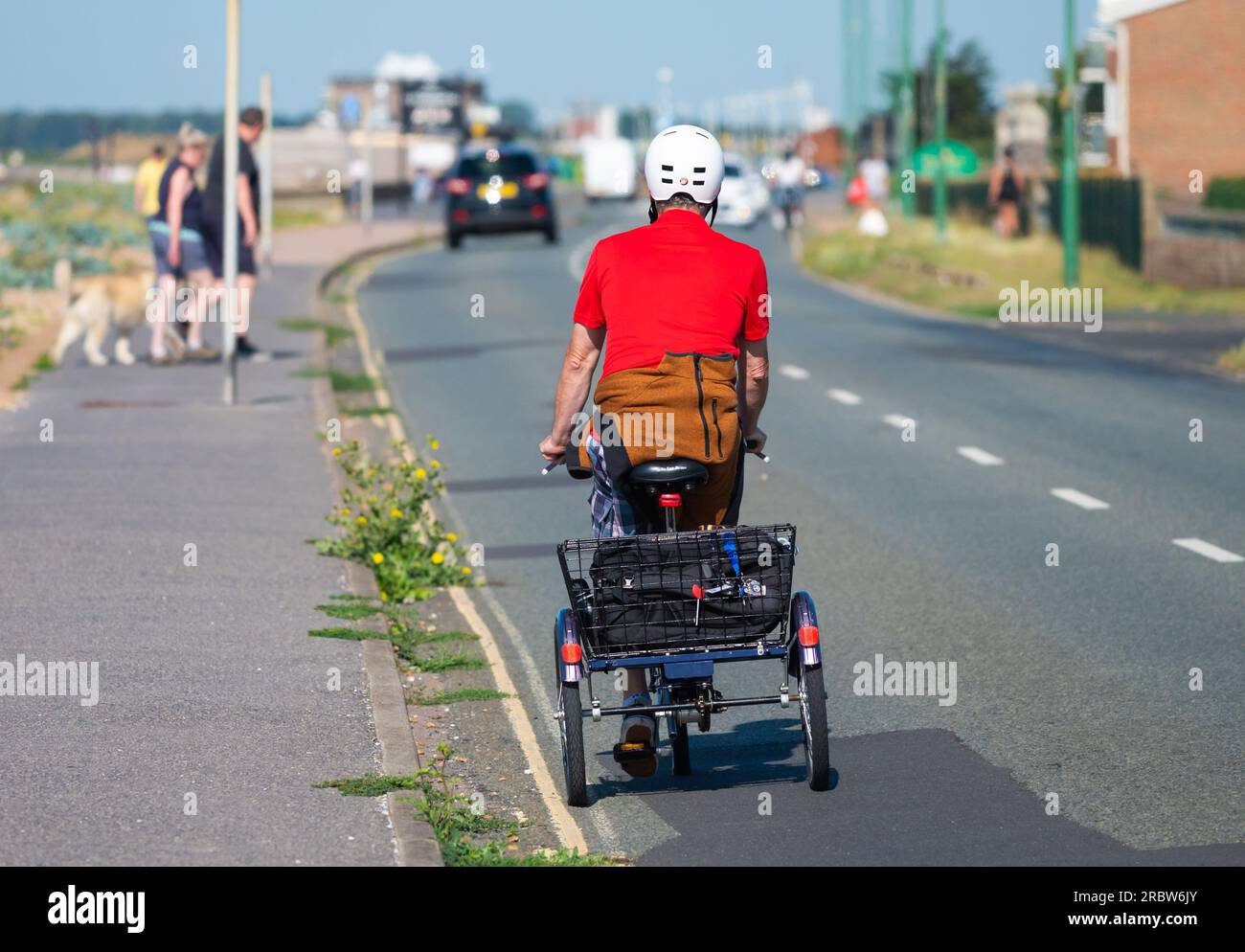Man riding tricycle, a pushbike with 3 wheels, with a shopping basket, wearing a helmet on a seafront road in Summer, UK. Stock Photo