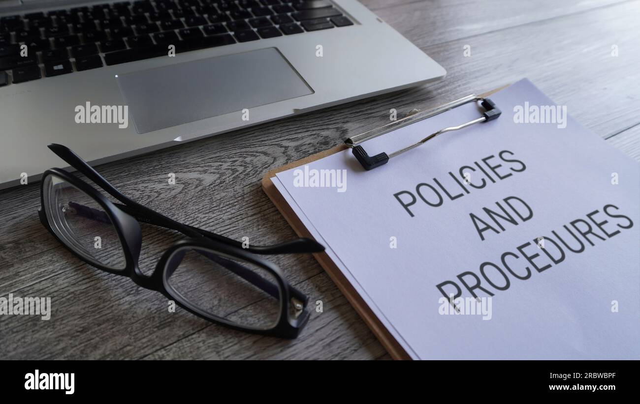 Paper clipboard with text POLICIES AND PROCEDURES on table with laptop and glasses. Stock Photo