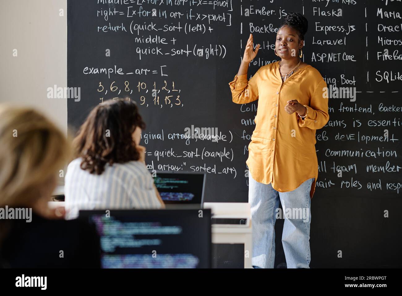 Teacher standing against the blackboard and teaching IT lesson to ...