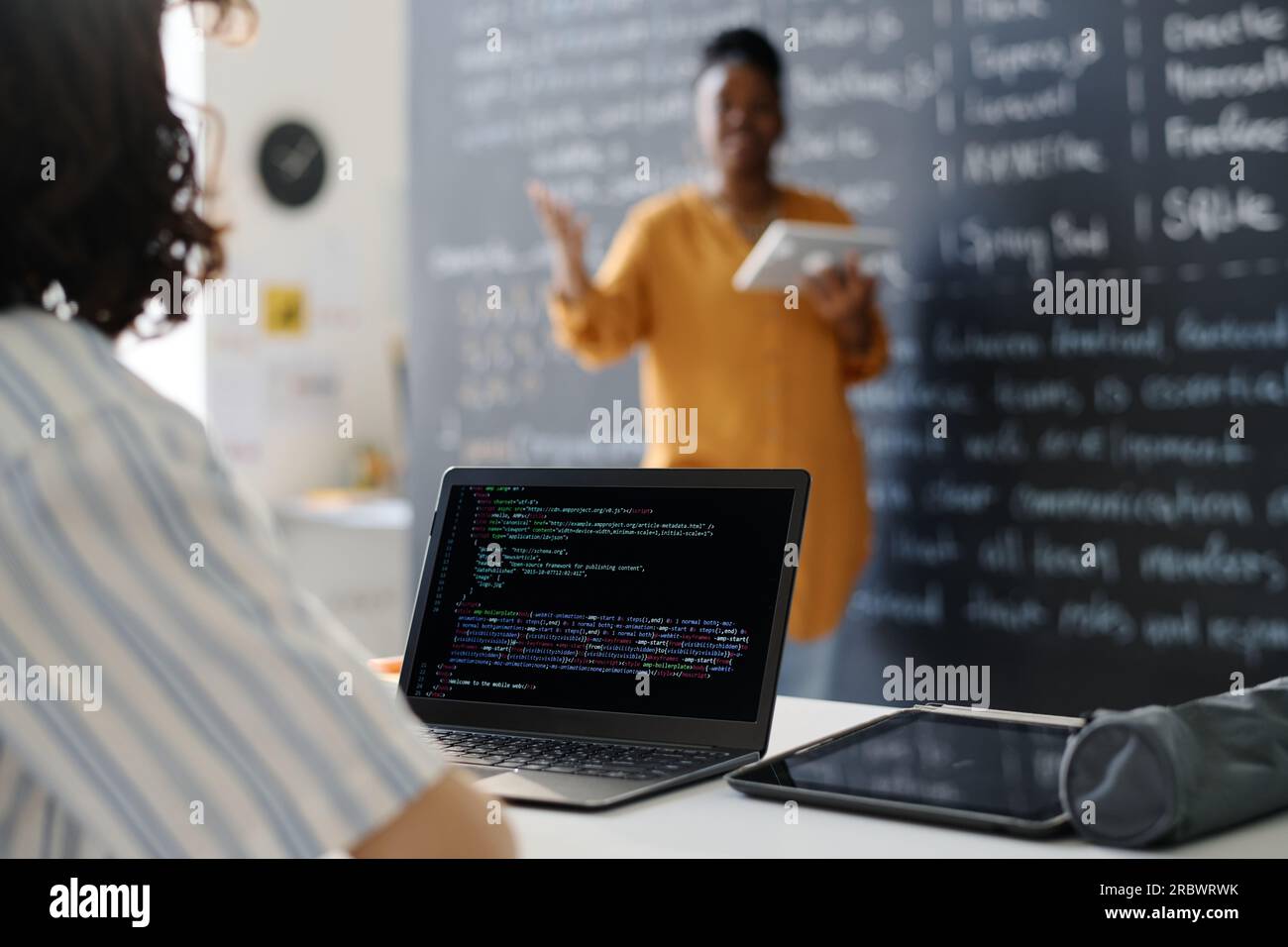 Rear view of student typing codes on computer while sitting at desk at lesson with teacher explaining material in background Stock Photo