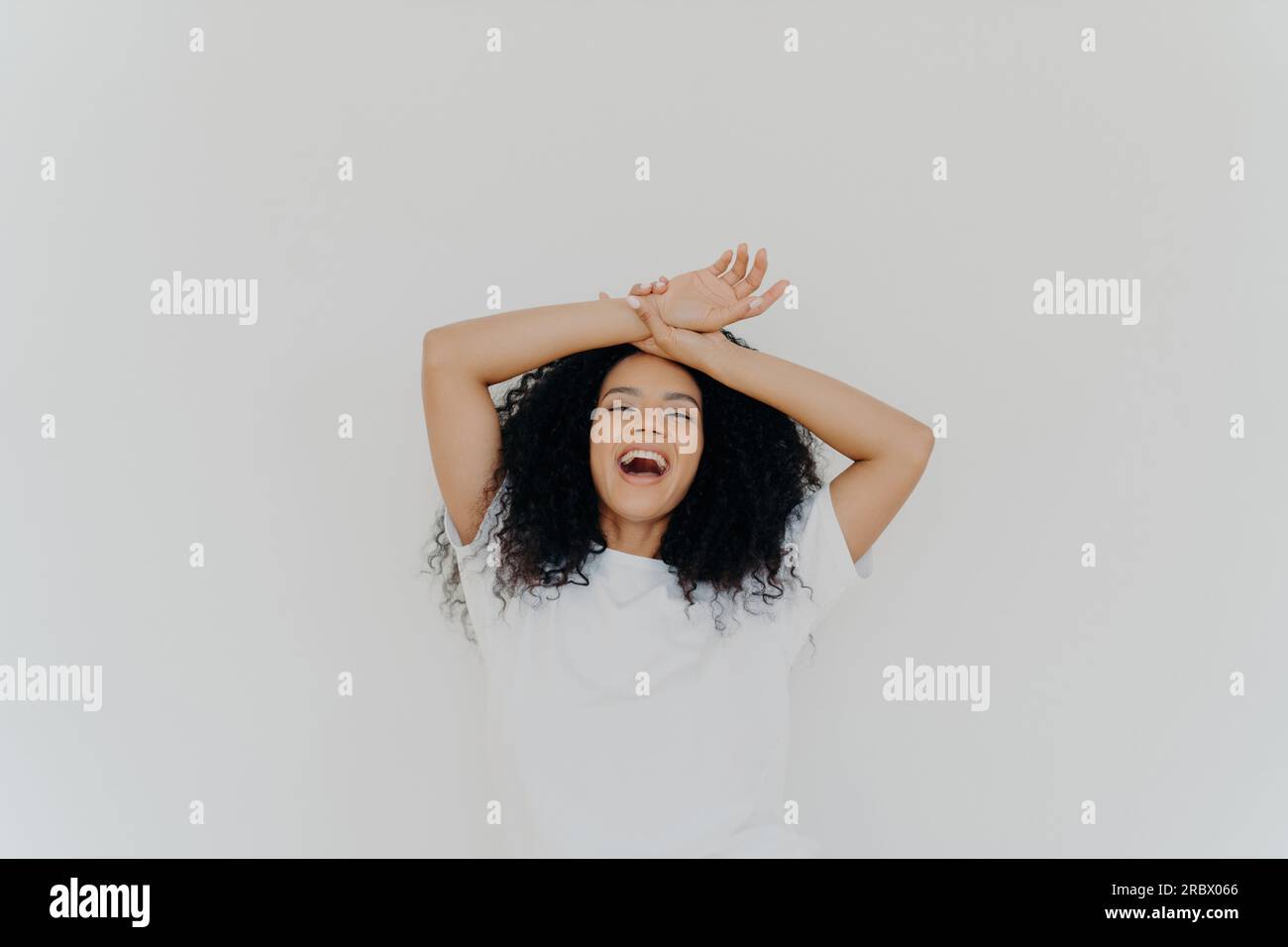 Overjoyed curly-haired lady, hands on forehead, laughing with mouth wide open, energetic vibes. Casual attire, white studio backdrop. Hilarious joke! Stock Photo