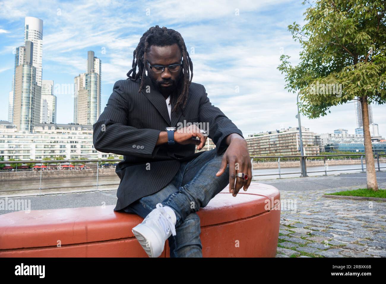 young African business man with beard, dreadlocks and glasses, in casual clothes, wears a blue jacket, is waiting outdoors, sitting watching the time, Stock Photo