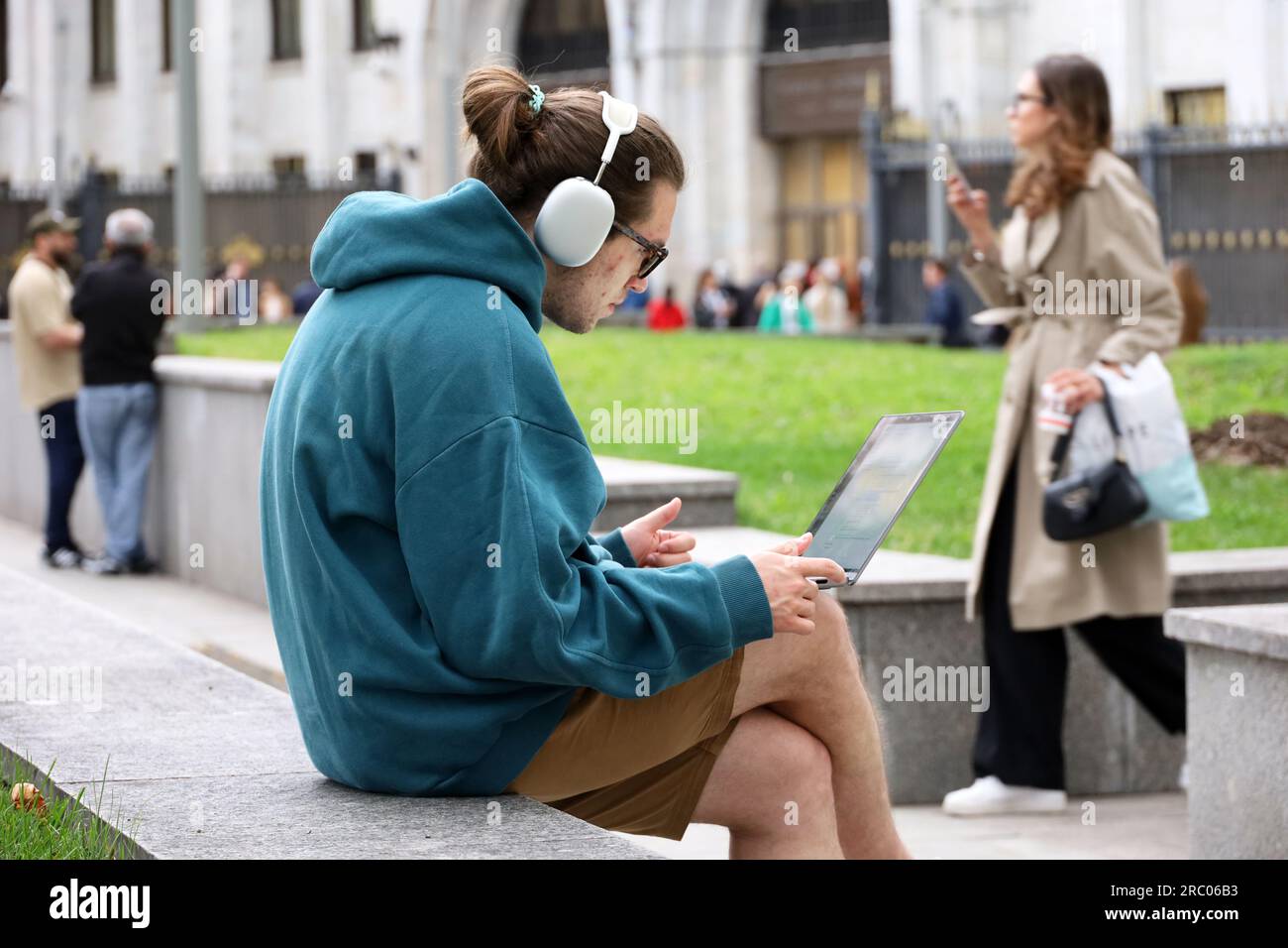 Guy in shorts and headphone sitting with a laptop on a bench on summer street Stock Photo