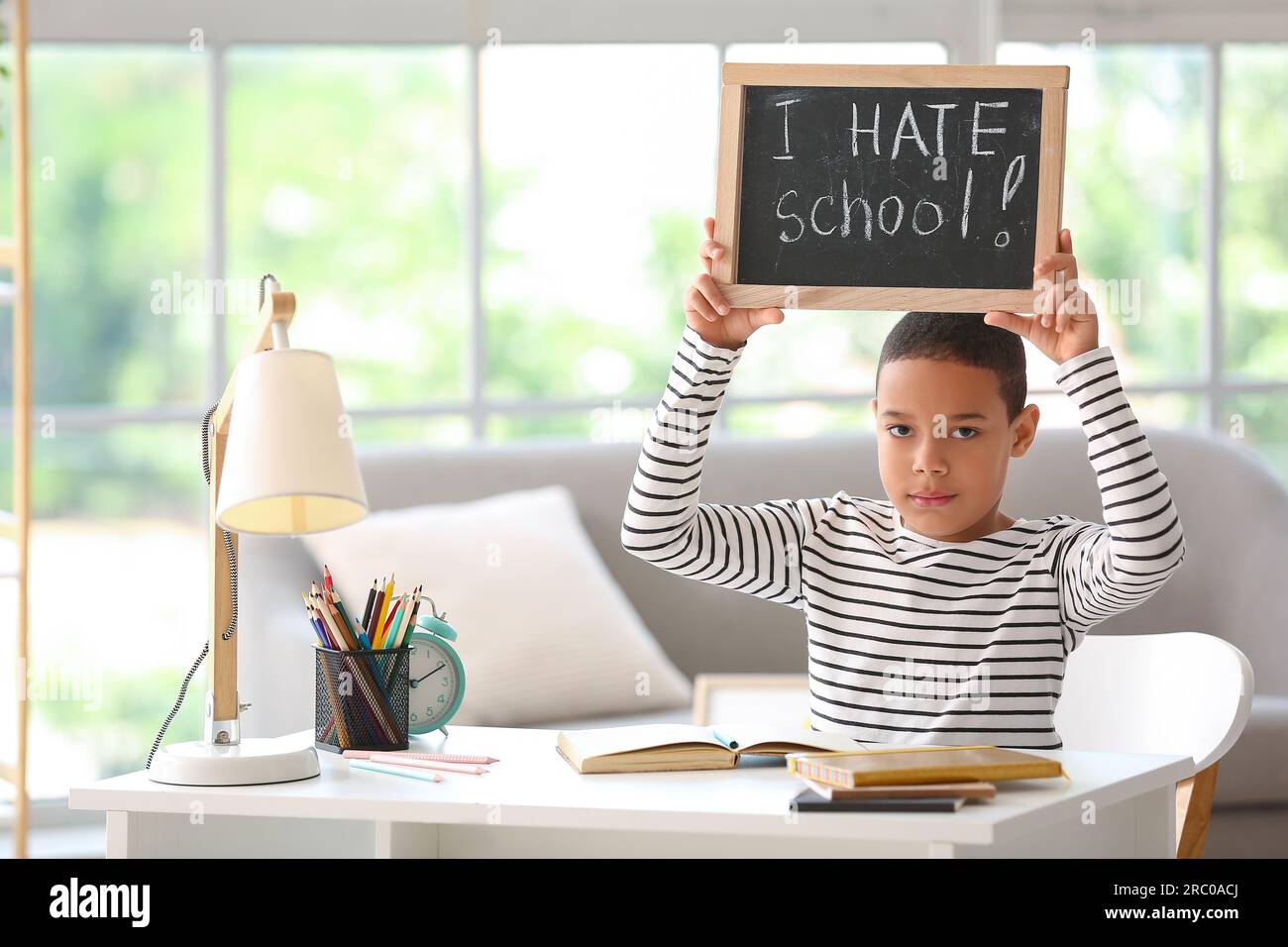 Little African-American boy holding chalkboard with text I HATE SCHOOL at home Stock Photo