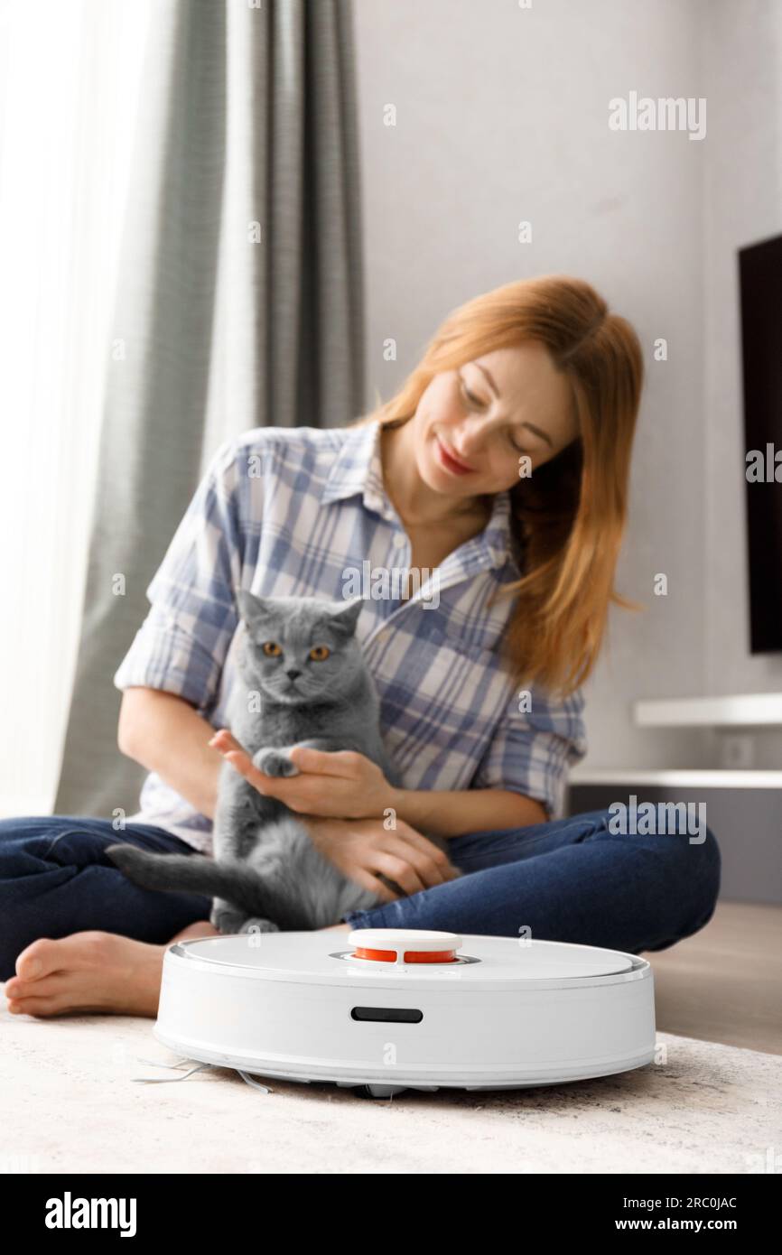 A smart robot vacuum cleaner cleans the carpet, a girl and a cat look at the vacuum cleaner. The concept of smart home, daily cleaning Stock Photo