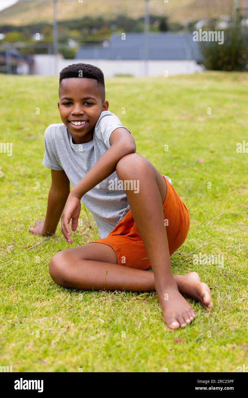Portrait of happy african american schoolboy sitting on grass at school ...