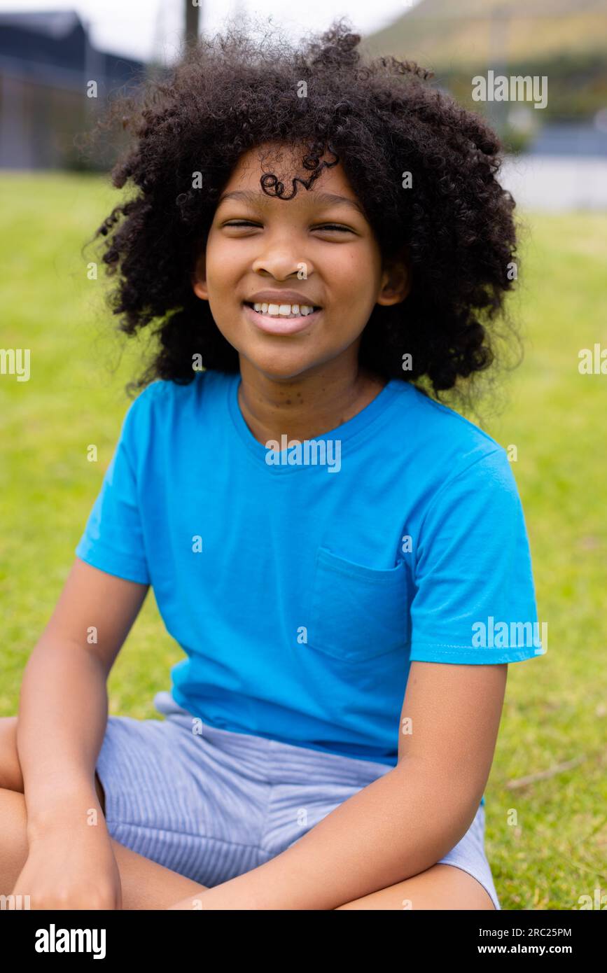 Portrait of happy african american schoolboy sitting on grass at school ...