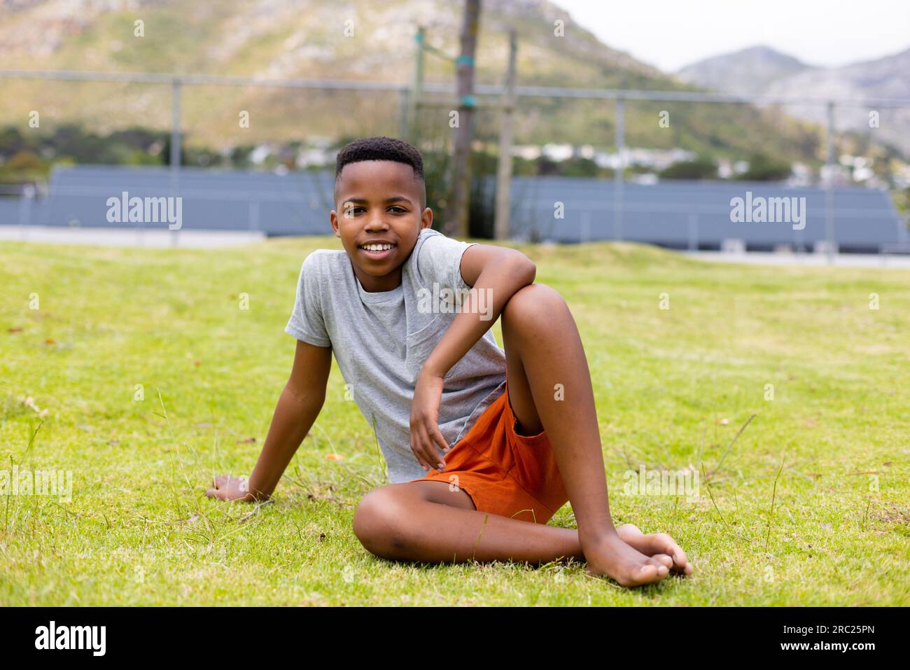 Portrait of happy african american schoolboy sitting on grass at school ...