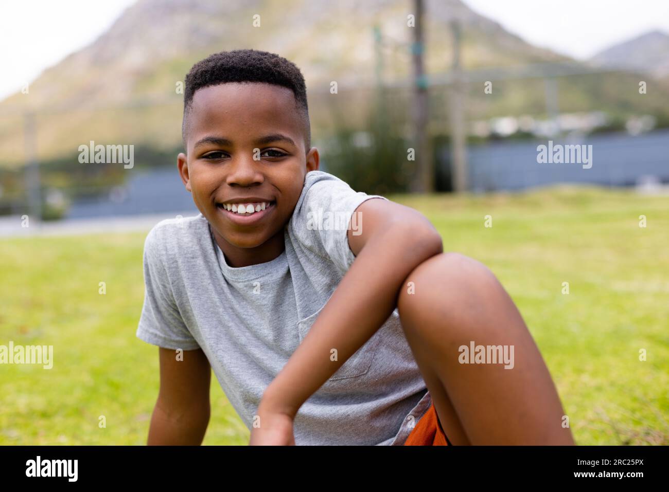 Portrait of happy african american schoolboy sitting on grass at school ...