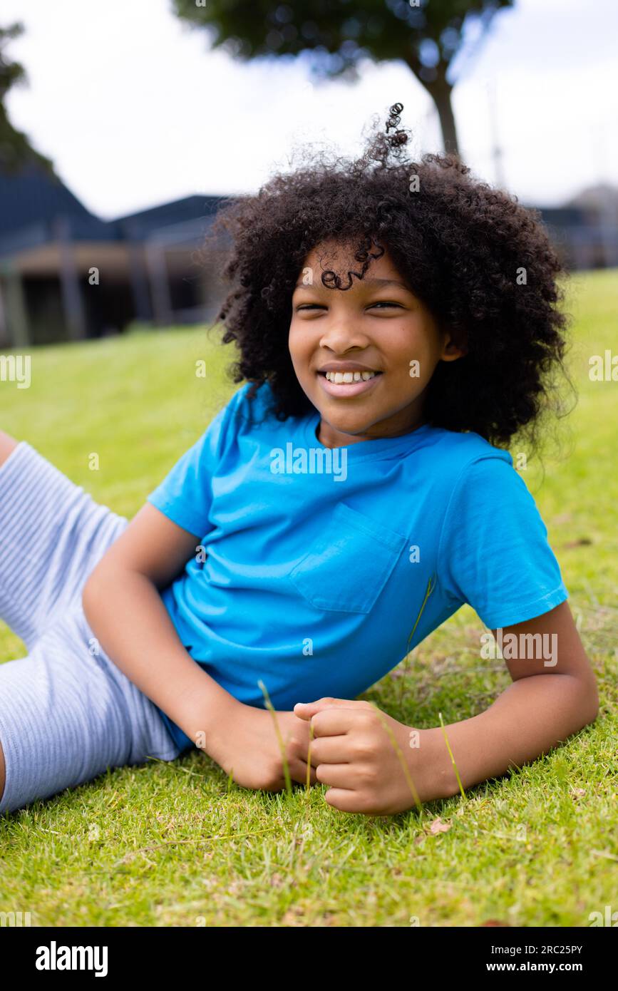 Portrait of happy african american schoolboy sitting on grass at school ...