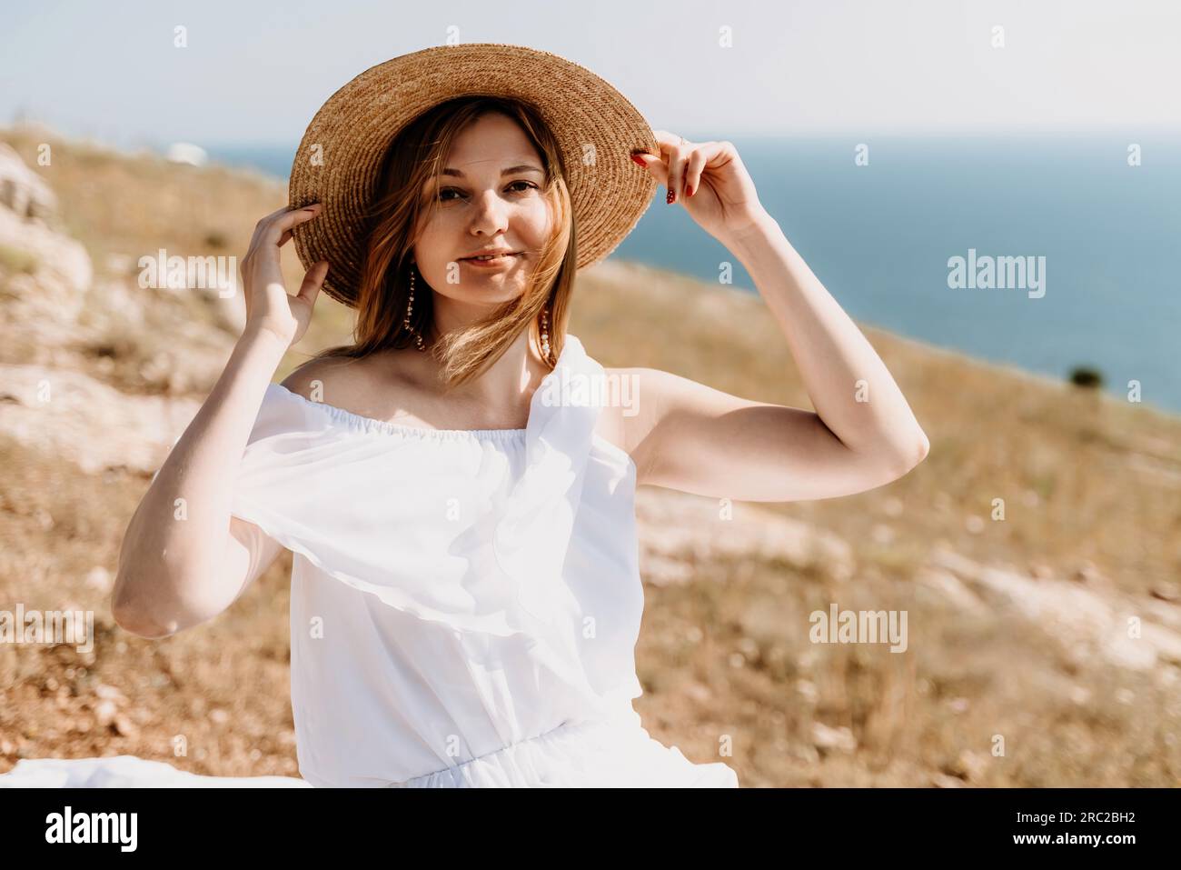 Happy woman in a white dress and hat stands on a rocky cliff above the ...