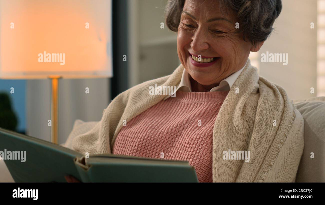Happy smiling laughing Caucasian old woman reading bestseller book peace at home covered with blanket senior mature lady smile elderly female retired Stock Photo