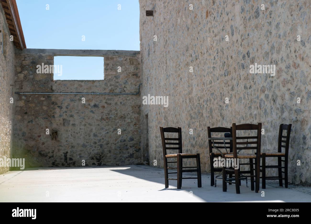 Four empty wooden wicker chair in shadow, stonewall around with open roof. Elefsina, Attica Greece. Mind freedom. Under view of blue sky. Stock Photo