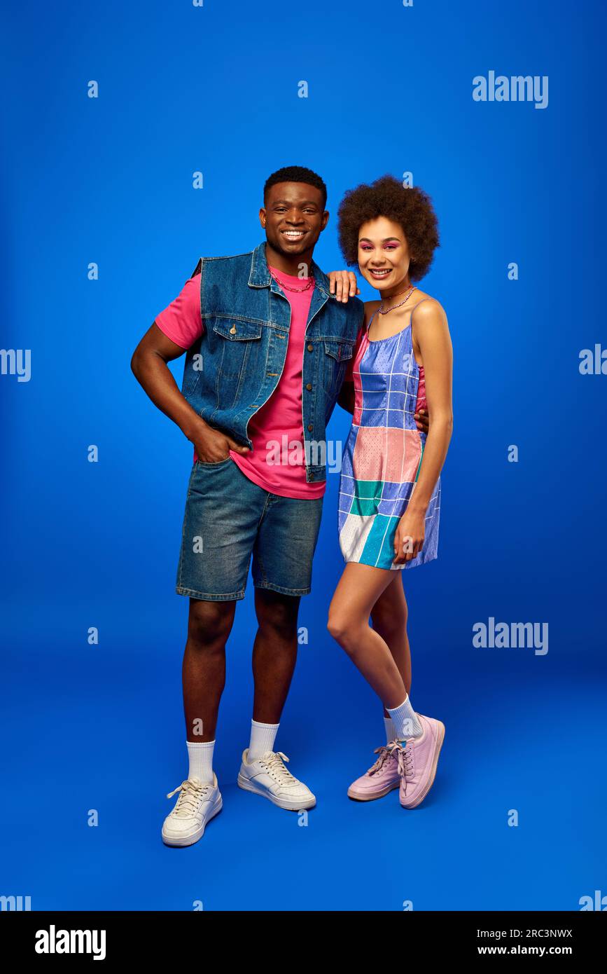 Full length of trendy and cheerful young african american best friends in summer clothes posing and looking at camera while standing on blue backgroun Stock Photo