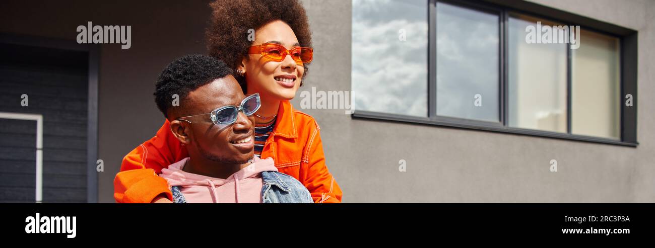 Positive young african american woman in sunglasses and modern outfit hugging best friend and looking away while standing together near building on ur Stock Photo