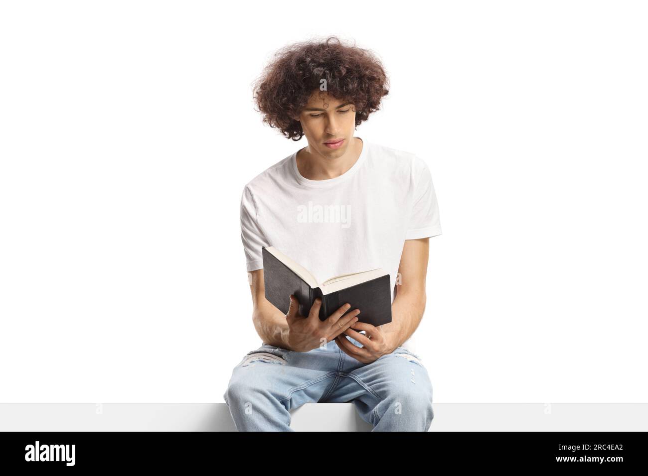 Young man with a curly hair sitting on a panel and reading a book isolated on white background Stock Photo