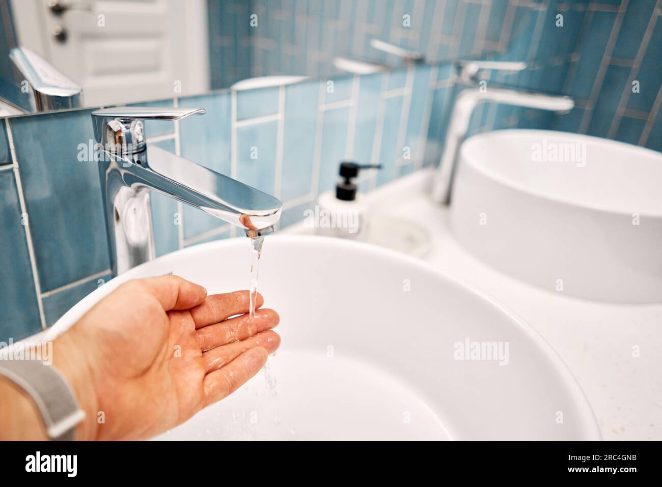 A man's hand touches the water in a beautiful sink with a metal faucet next to an mirror. Close-up of an elegant faucet in the bathroom sink next to s Stock Photo