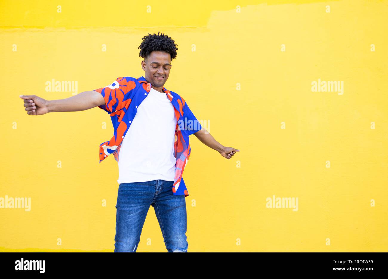 Cheerful American male with dreadlocks and in colorful shirt dancing while swinging hands against yellow background Stock Photo