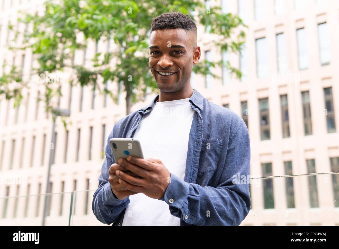 Smiling African American male entrepreneur in casual outfit smiling and text messaging on mobile phone while working on city street and looking at cam Stock Photo