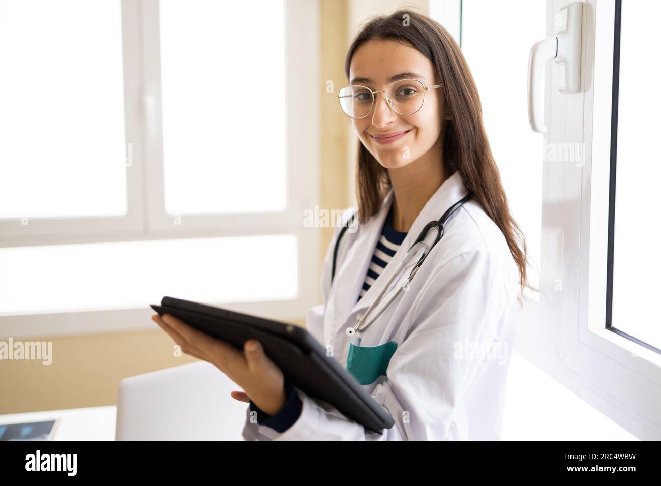 Young female doctor in white coat and eyeglasses looking at camera of tablet while standing in office of modern clinic Stock Photo