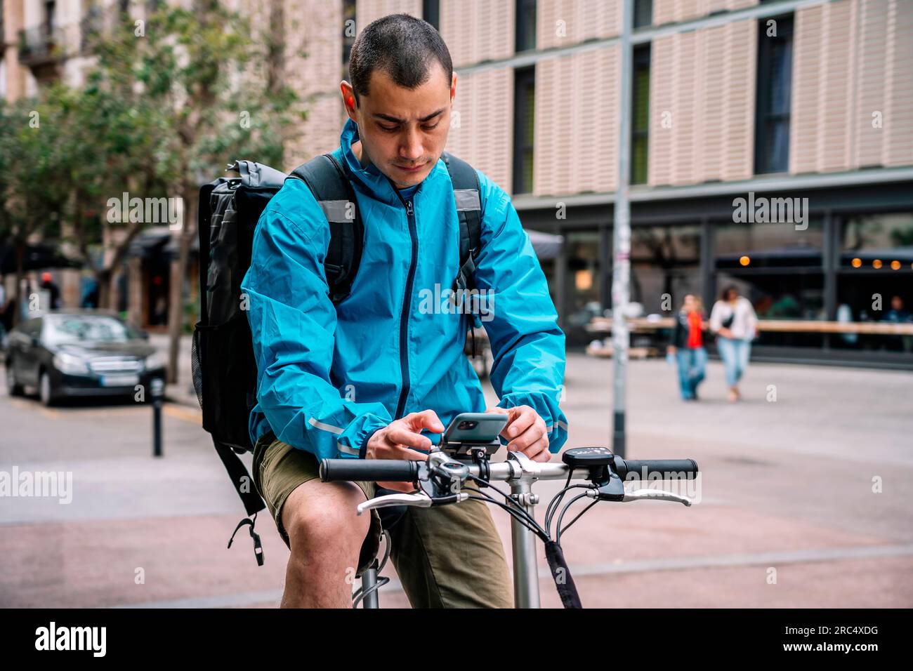 Delivery man in bright blue jacket with backpack sitting on bicycle in city street and using phone while working in Barcelona Stock Photo